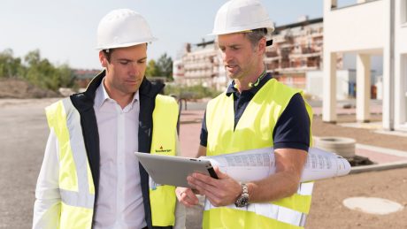 Two men with safety vests; white helmets, holding a tablet, building site