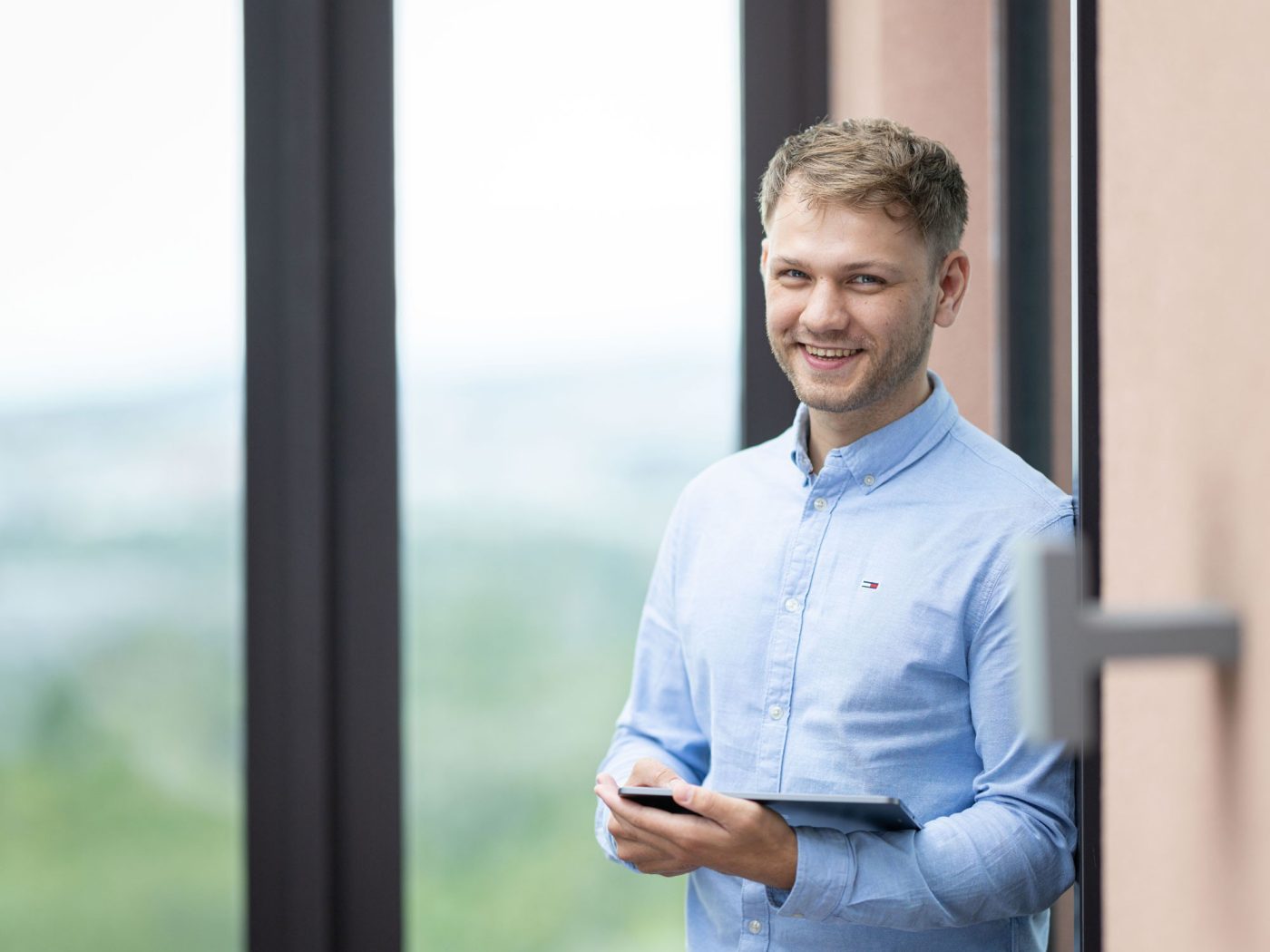 Ein Mann hält ein Tablet, lehnt an einer Wand und lächelt in die Kamera