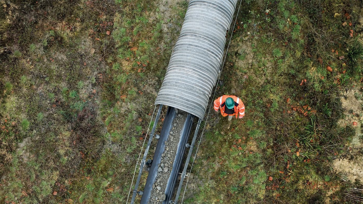 Vogelperspektive, Mann mit oranger Sicherheitsjacke und grünem Helm geht neben einem Förderband für Rohmaterial in der Natur