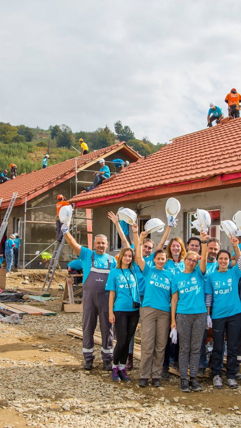 Menschen mit blauen T-Shirts halten weiße Helme in die Luft, Rohbauhäuser im Hintergrund