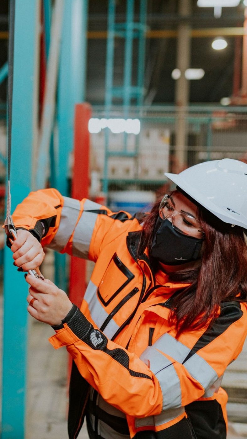 Frau hilft Mann Schutzausrüstung anzulegen, Produktionshalle von innen