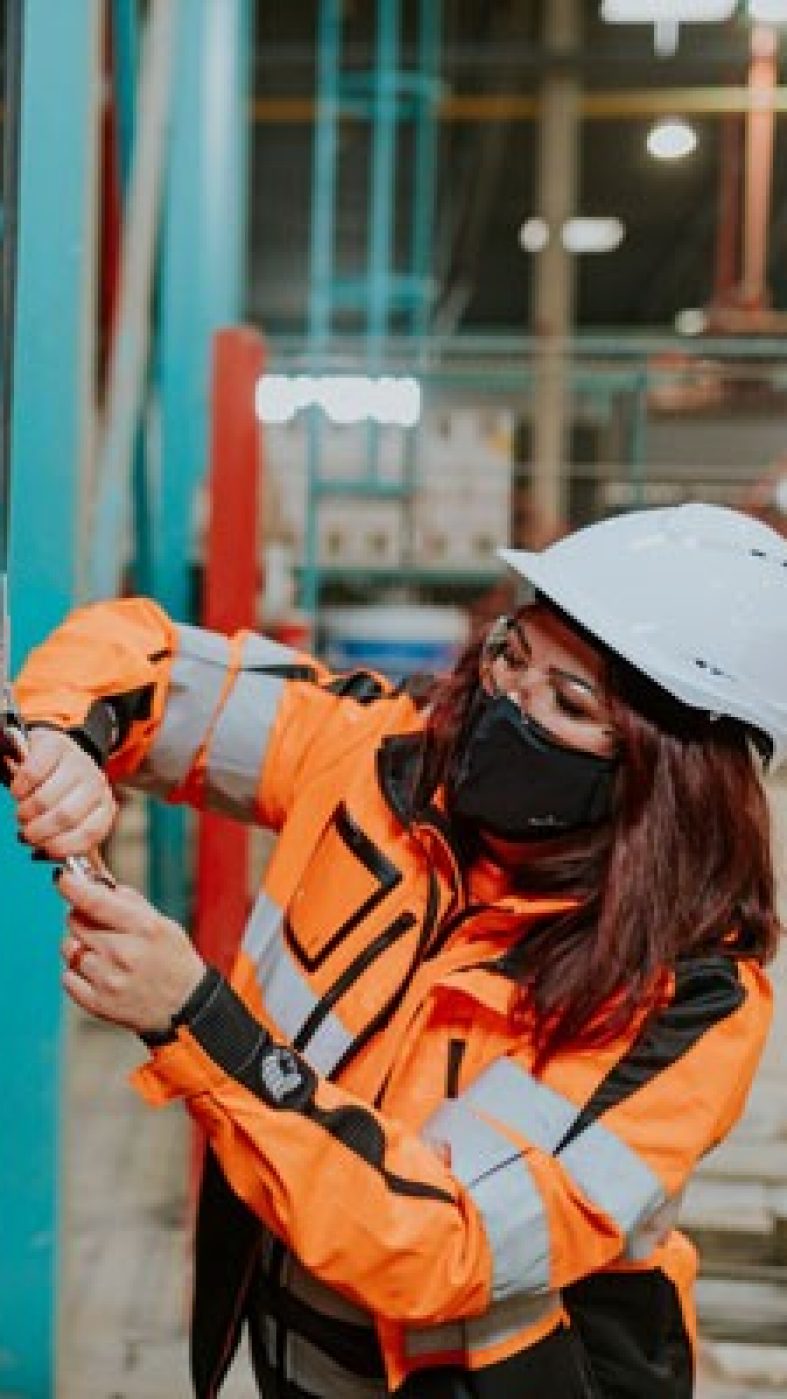 Frau hilft Mann Schutzausrüstung anzulegen, Produktionshalle von innen