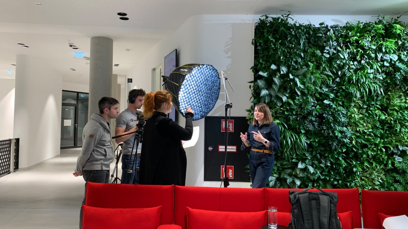 Videodreh, vier Personen, Frau spricht, rotes Sofa, Pflanzenwand