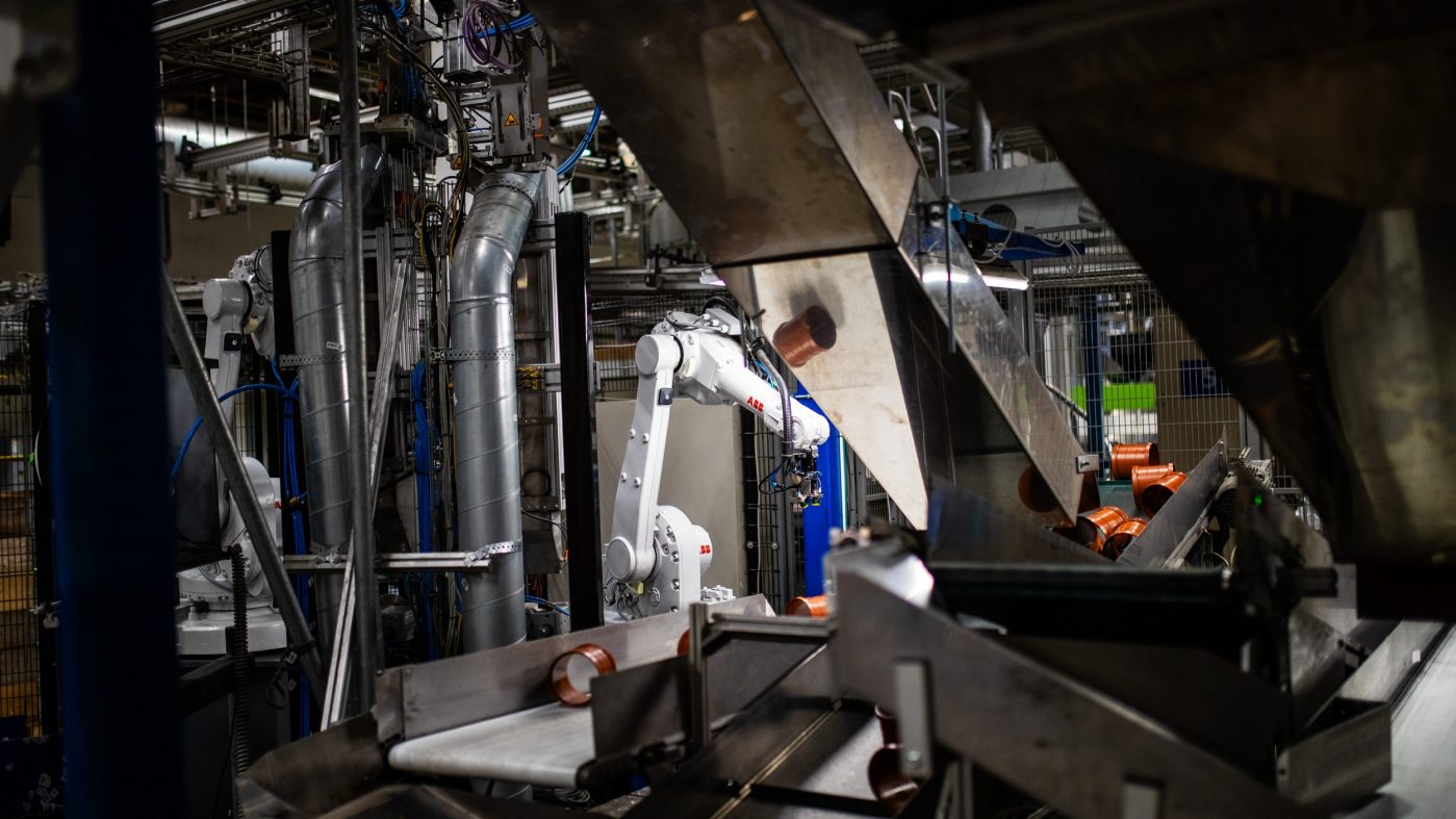 Ein Roboterarm arbeitet an einer Produktionslinie von Pipelife, umgeben von Metallrohren, Förderbändern und Industrieanlagen.