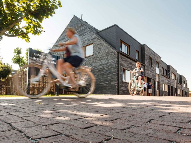 Woman and child riding bike over street of cobblestones, blurred, brick house