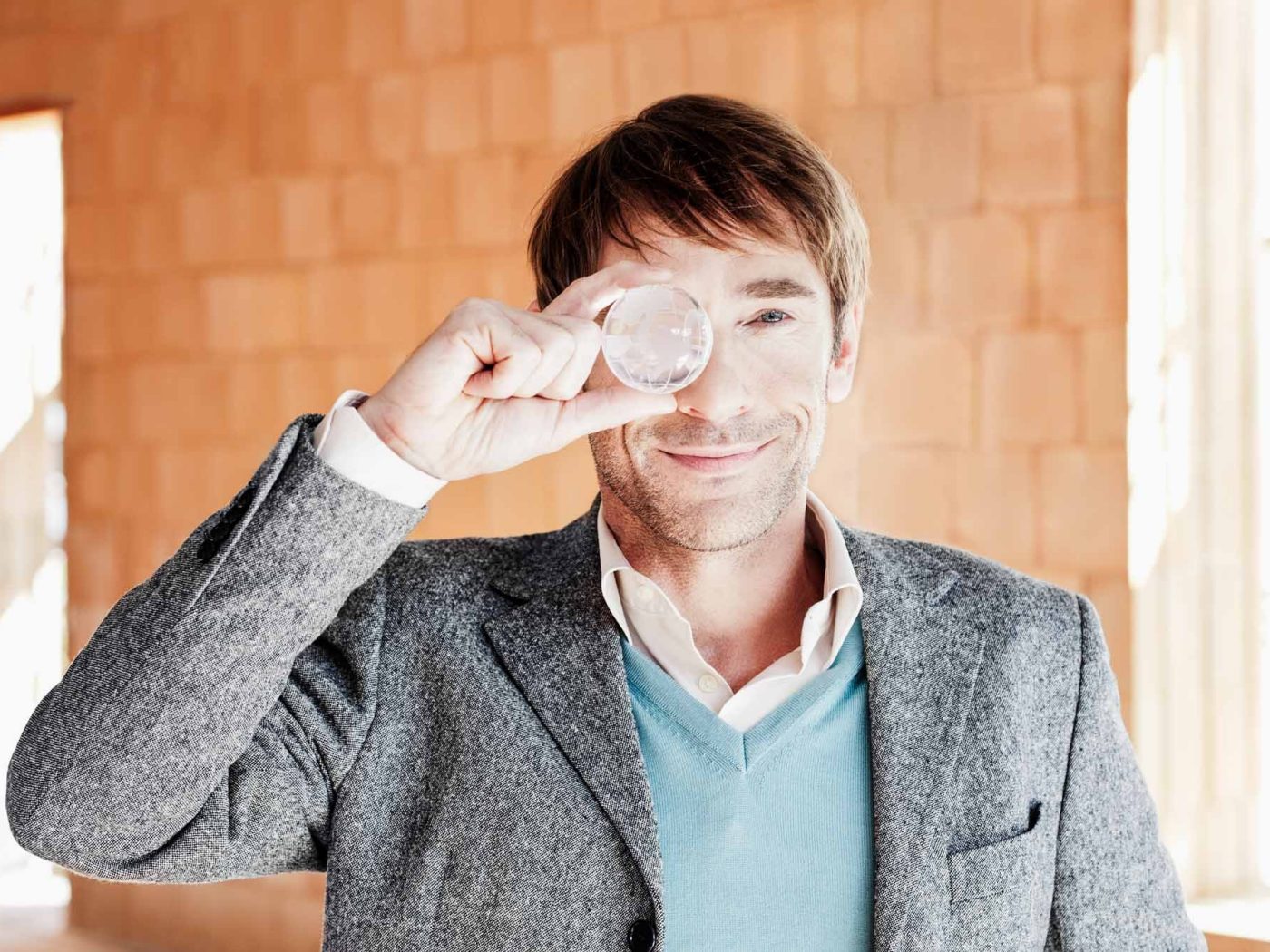 Men holding a glas globe in front of his right eye in front of a brick wall