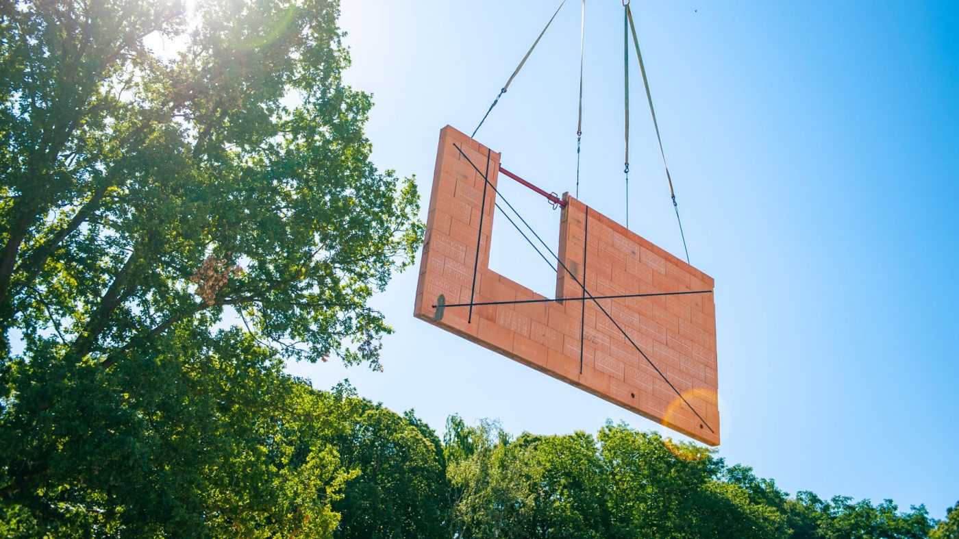 Fertigteilwand wird von einem Krahn hochgehoben, blauer Himmel und grüne Bäume im Hintergrund