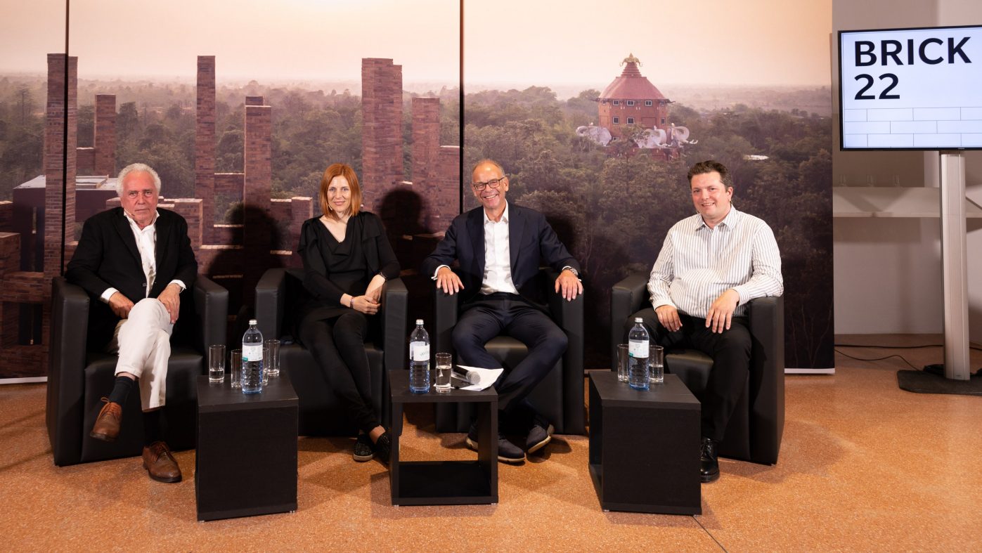 Panel discussion, four people sitting on black armchairs and looking into the camera