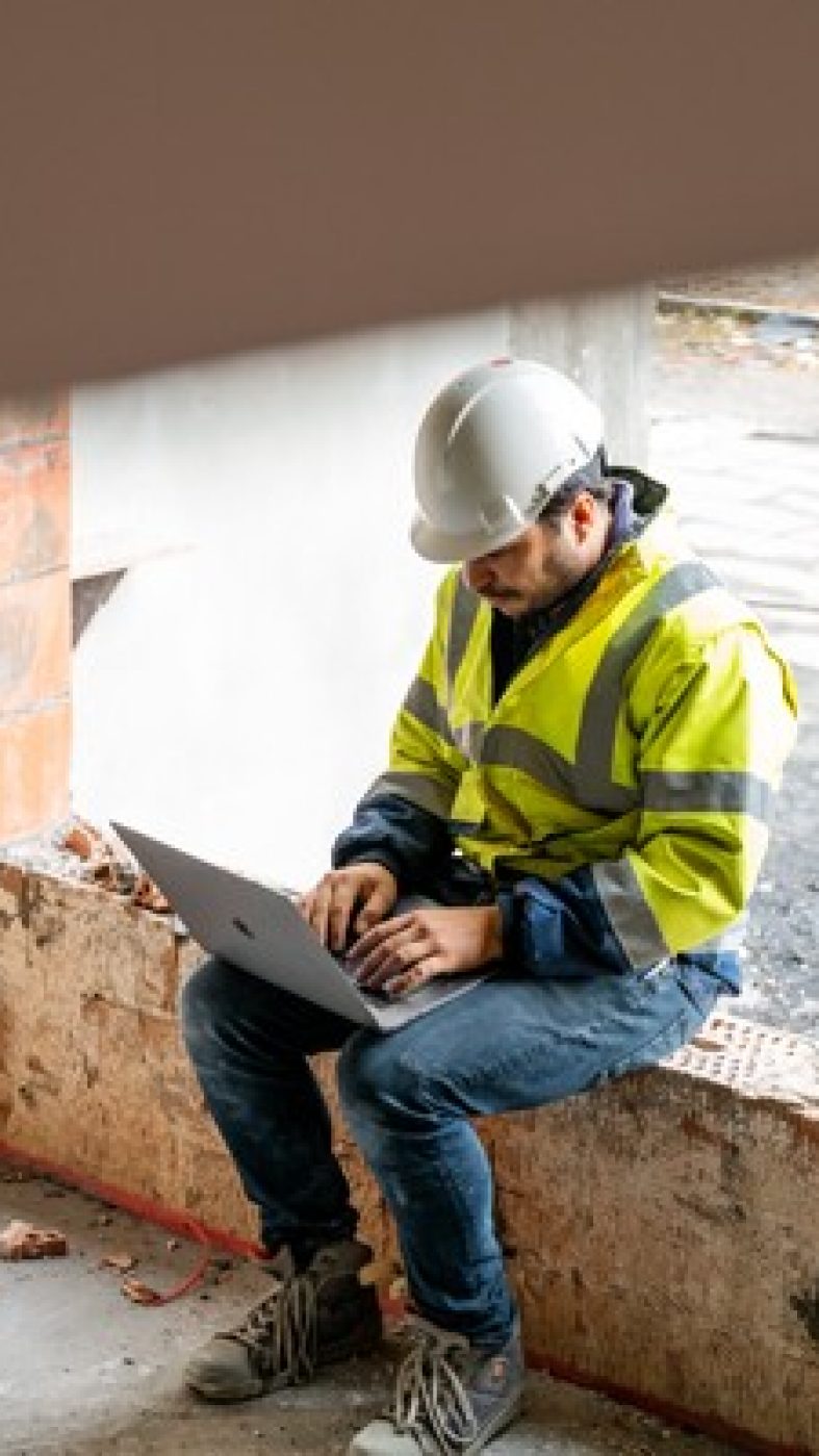 Man sitting on a window sill in a raw building with a laptop in protective clothing