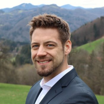 Man grinning, suit, on green meadow, mountains in background