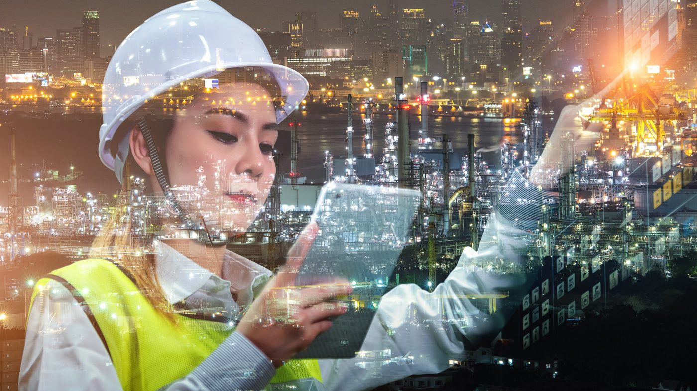 Working in Industry 4.0: An engineer in a hard hat and safety jacket with a tablet against the background of an illuminated factory.