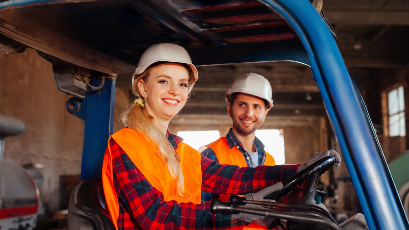 Portrait of smiling woman, forklift driver sitting in a truck, looking at the camera. Young man, warehouse worker looking with admiration at his female colleague, forklift driver.