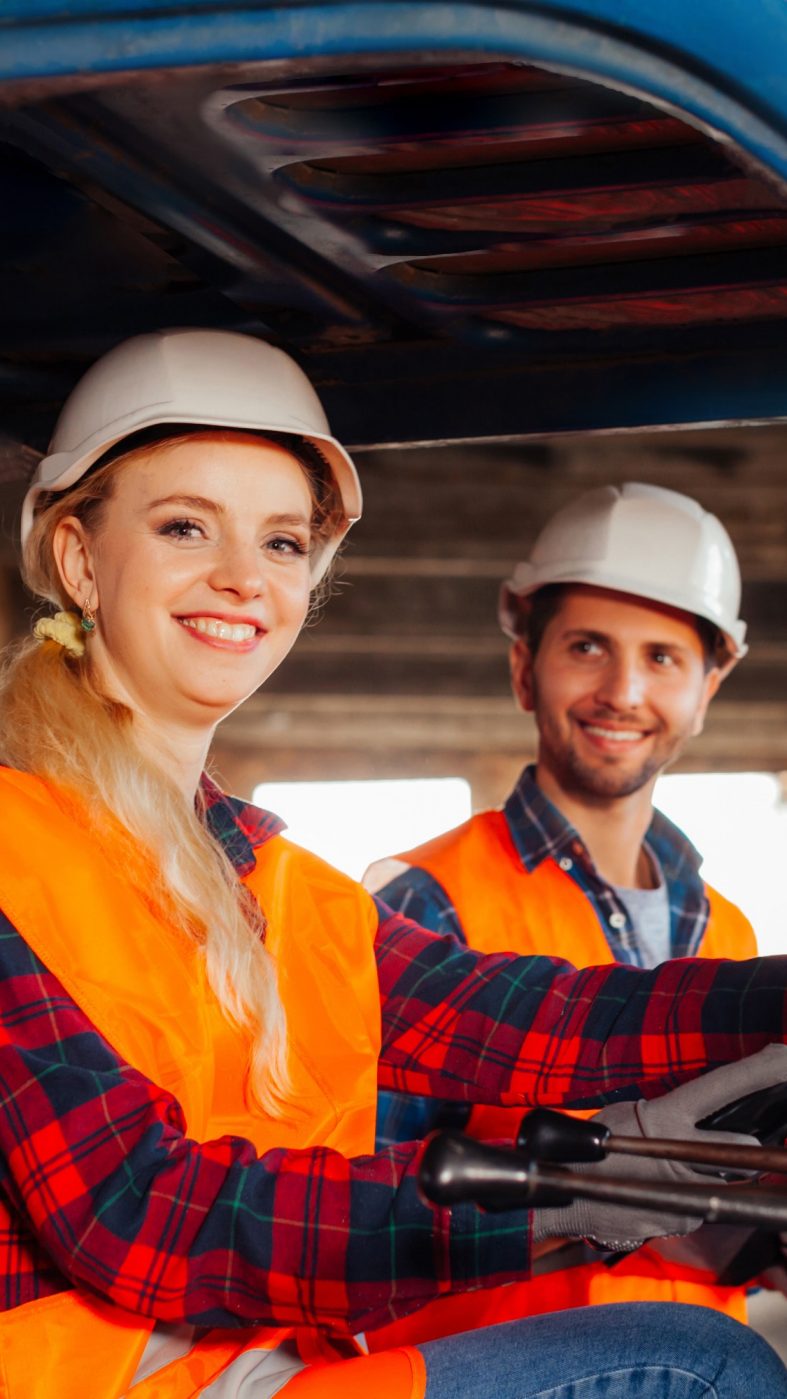 Portrait of smiling woman, forklift driver sitting in a truck, looking at the camera. Young man, warehouse worker looking with admiration at his female colleague, forklift driver.