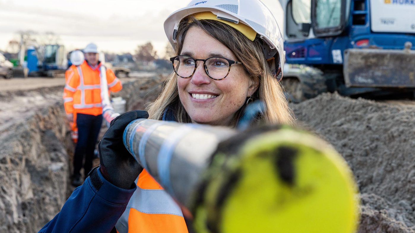 Woman with glasses and helmet carries a pipe on her shoulder and smiles