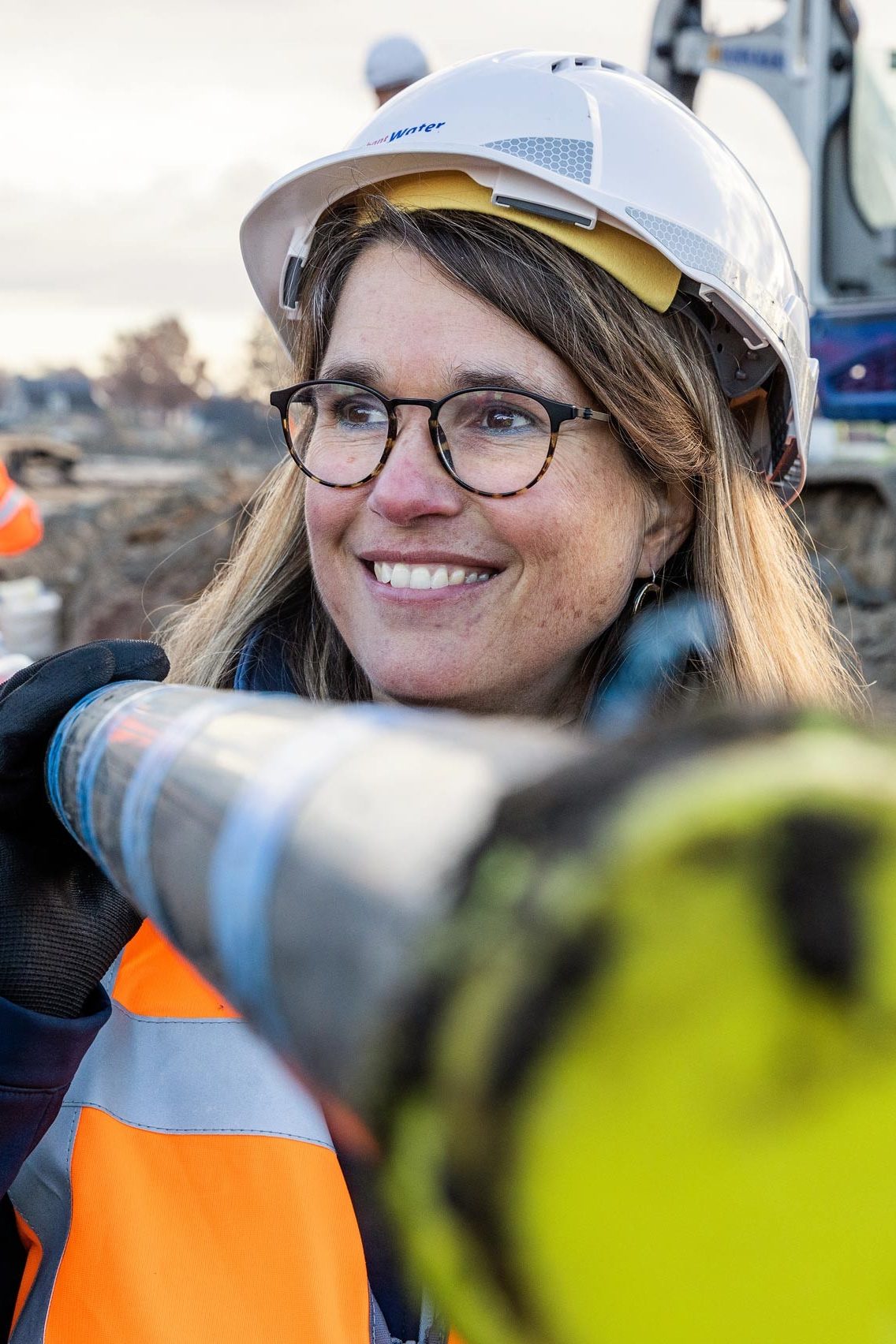 Woman with glasses and helmet carries a pipe on her shoulder and smiles