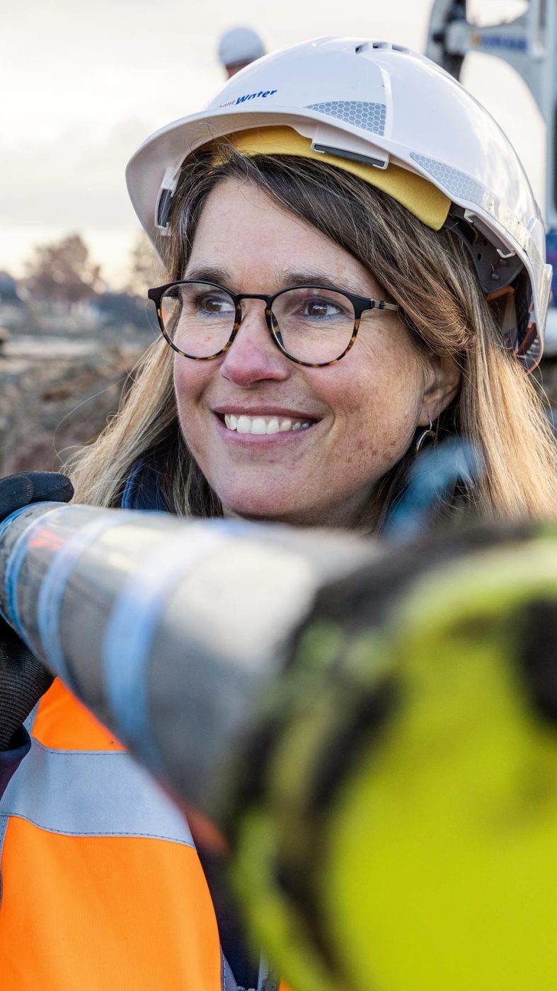 Woman with glasses and helmet carries a pipe on her shoulder and smiles