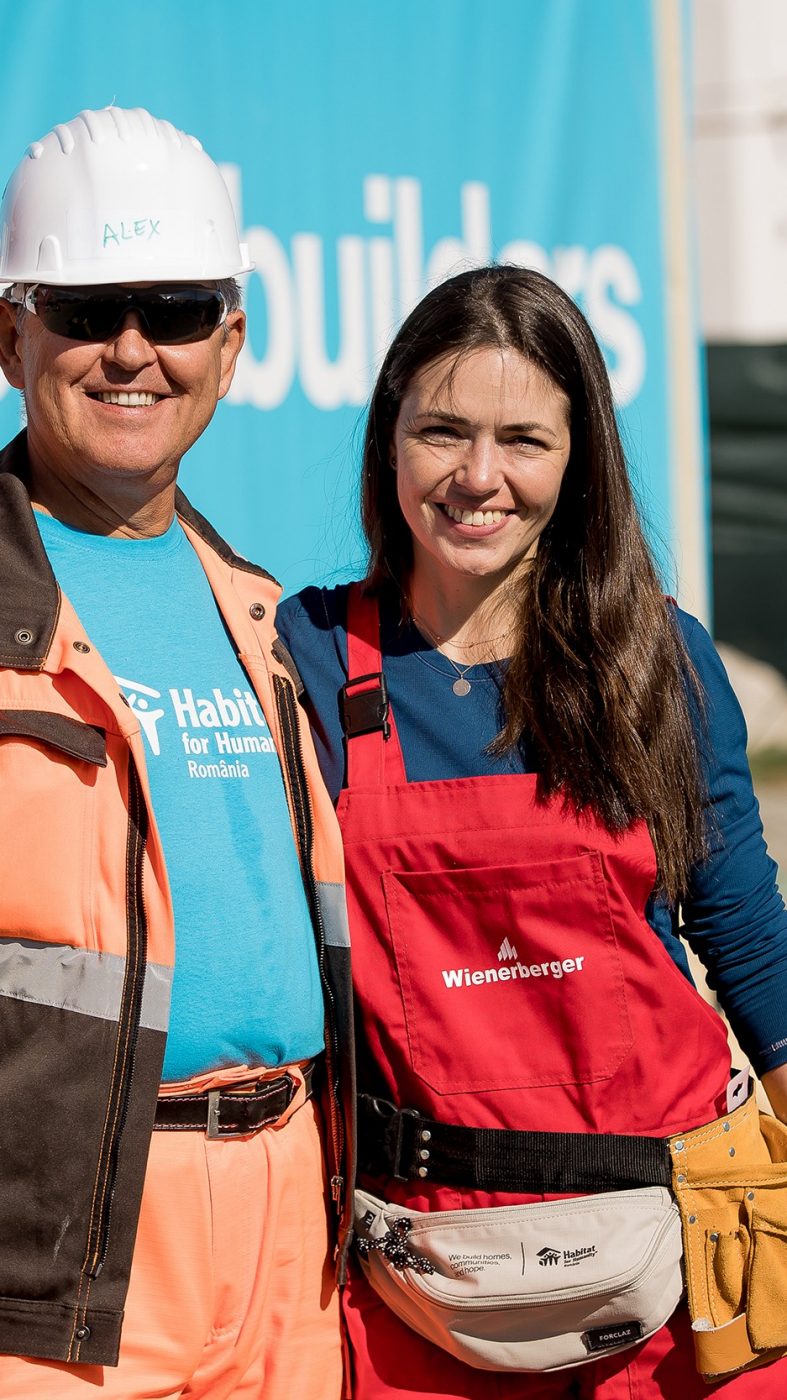 a man and a woman in work clothes laugh into the camera in front of a construction site